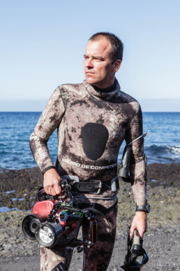 Una persona con un traje de neopreno de camuflaje se encuentra de pie junto al océano, sosteniendo un equipo de buceo y una cámara submarina lista para usar en editoriales. El cielo está despejado y la costa rocosa se ve al fondo.