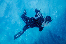 Una fotografía editorial capta a un buceador con traje de neopreno, nadando bajo el agua con una cámara de gran tamaño. El agua azul vibrante y las burbujas que la rodean crean una escena acuática envolvente, mientras el buceador, equipado con aletas, hace un gesto con una mano.