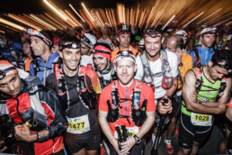 Un grupo de corredores de trail con dorsales numerados y linternas frontales se reúne en la línea de salida de la carrera, destilando entusiasmo y disposición. Al fondo, otros corredores contribuyen a la atmósfera animada, creando una escena enérgica perfecta para un editorial que captura la esencia de la competencia.