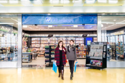 Dos mujeres, con estilo editorial, sonríen al salir de una tienda Hudson, cada una con una bolsa de compras azul en la mano. La entrada está repleta de revistas y artículos diversos. Un suelo amarillo complementa el ambiente luminoso y acogedor.