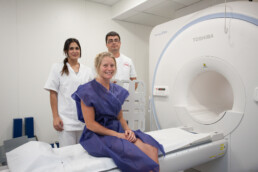 Una mujer con bata médica está sentada en la camilla de una máquina de resonancia magnética y sonríe a la cámara en un entorno de estilo editorial. Dos profesionales de la salud, un hombre y una mujer, posan detrás de ella en la moderna sala de imágenes médicas.