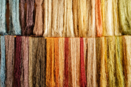 Los manojos de hilo multicolor cuelgan verticalmente como si fueran un editorial, mostrando un degradado de tonos cálidos que van desde marrones y ocres hasta dorados, naranjas y rojos apagados. La textura fibrosa agrega un acabado ligeramente áspero a la vibrante exhibición.