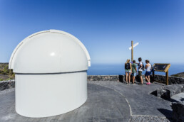 Un grupo de personas se encuentra de pie en un sendero de piedra cerca de una cúpula blanca de observatorio, participando en una exploración editorial mientras se reúnen alrededor de un cartel informativo y un poste direccional. El cielo despejado les ofrece una vista sin obstáculos del océano al fondo.