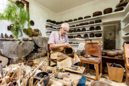 En un entorno digno de un editorial, un hombre mayor trabaja meticulosamente en un taller de cerámica. Está rodeado de vasijas terminadas en estanterías y de varias herramientas sobre la mesa. La habitación es rústica, con paredes de piedra y un helecho que aporta un toque de verdor.