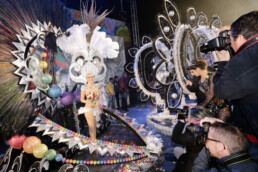 Una artista con un elaborado y colorido traje con plumas y destellos posa en un escenario. Fotógrafos editoriales la rodean, capturando el momento en medio de un vibrante telón de fondo y una atmósfera festiva.