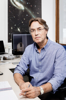 Un hombre con gafas y camisa azul está sentado en un escritorio en una oficina. Sostiene un bolígrafo sobre un cuaderno abierto, listo para redactar un artículo editorial. Cerca, los ordenadores zumban y un póster cosmológico añade profundidad al espacio de trabajo creativo que se ve al fondo.