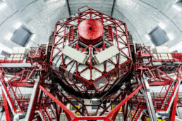 Un primer plano editorial muestra un instrumento científico grande y complejo con un marco de metal rojo y numerosos paneles, ubicado dentro de una instalación espaciosa de estilo industrial con un techo alto y curvo.