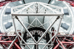 La vista interior de un gran telescopio muestra una red de soportes y vigas de metal blanco y negro. Se ven elementos estructurales rojos en los laterales, mientras que el techo abovedado, adornado con iluminación industrial, crea un aspecto brillante y futurista.