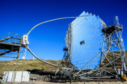 Un gran telescopio circular con una superficie espejada se alza contra un cielo azul claro y capta un paisaje digno de un editorial. Apoyado sobre una estructura metálica con escaleras, domina colinas bajas y vegetación escasa, creando un paisaje propicio para la exploración.