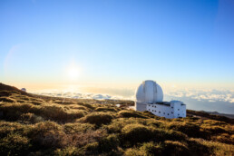 Una cúpula de observatorio de un blanco prístino reposa sobre una colina iluminada por el sol y con una exuberante vegetación, dominando majestuosamente un mar de nubes bajo un cielo azul claro. En una escena digna de un editorial, el sol proyecta su cálido resplandor al salir o ponerse en el horizonte, tiñendo el paisaje de tonos dorados.