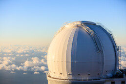 Una gran cúpula blanca de observatorio con el techo abierto se yergue sobre las nubes al atardecer, bañada por un encanto editorial. El cielo azul suave forma su telón de fondo, mientras una escalera y una barandilla de metal envuelven con gracia su forma esférica.