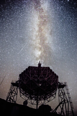 La silueta de una gran estructura intrincada de telescopio se destaca en el cielo nocturno estrellado. La Vía Láctea forma una banda brillante a lo largo de la escena, con estrellas densamente dispersas alrededor. En la parte superior izquierda, una débil estrella fugaz agrega un toque de asombro a la composición.
