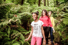 Tres personas caminan por un frondoso sendero forestal bordeado de helechos en una escena digna de un editorial. La persona que va delante lleva una camiseta blanca con texto y pantalones rojos, seguida por otras personas con camisetas de colores rojo y amarillo vibrantes, que capturan la esencia de la exploración de la naturaleza.