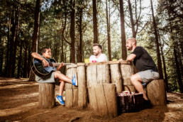 Tres personas están sentadas sobre troncos de madera alrededor de una mesa de troncos en un bosque, creando una escena digna de un editorial. Una toca la guitarra mientras los demás escuchan con sonrisas. Hay una canasta de picnic cerca y la luz del sol se filtra hermosamente a través de los árboles.