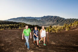 Tres personas recorren un sendero de tierra digno de un artículo de opinión, rodeado de vegetación y montañas bajo un cielo azul claro. El grupo, formado por dos hombres y una mujer, incluye a una persona que sostiene un bastón mientras disfrutan del paisaje al aire libre.
