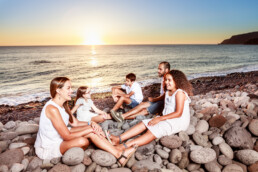Una familia de cinco personas se sienta en una playa rocosa durante la puesta de sol, creando un momento editorial. Los adultos y los niños, vestidos de blanco, se sonríen cálidamente. El mar en calma y el horizonte brillan con los tonos del sol poniente, capturando una noche perfecta.