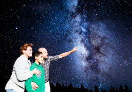 Una familia de tres personas se encuentra de pie bajo un cielo estrellado, mirando hacia arriba en una escena editorial. El hombre señala la Vía Láctea, que se extiende brillantemente a través de la noche. Las siluetas de los árboles se alinean en primer plano, realzando la belleza natural y la maravilla de la escena.