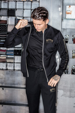 Una persona con un chándal negro de BOSS se encuentra de pie junto a una estantería de estilo editorial llena de artículos cuidadosamente ordenados, entre ellos corbatas y pequeñas cajas. Se ajusta ligeramente el cuello y mira hacia abajo.