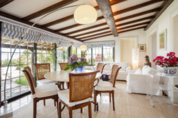Un solárium amplio y luminoso con grandes ventanales y vigas de madera ofrece espacios de relajación. Cuenta con una mesa de comedor redonda con sillas de madera, dos sofás blancos, flores de colores y una lámpara moderna. Toldos de rayas azules cubren las ventanas y dan a un exuberante jardín.