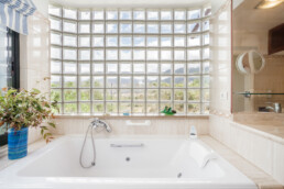 Un baño luminoso cuenta con una amplia bañera blanca en primer plano, situada debajo de una ventana de bloques de vidrio. La luz del sol se cuela a través de ella y ofrece una vista de la vegetación del exterior. Una planta en maceta se encuentra en la esquina de la bañera, lo que agrega un toque de naturaleza a los espacios de este entorno sereno.