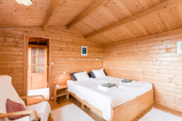 Un acogedor dormitorio tipo cabaña con paredes y techo de madera que captura la esencia de Espacios. Ofrece una cama doble con sábanas blancas, dos toallas cuidadosamente dobladas, una pequeña mesita de noche de madera y una silla acolchada. Una puerta de madera y un marco de fotos completan este acogedor refugio.