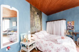 Un dormitorio acogedor cuenta con una colcha con motivos florales, un gran espejo en una pared azul y un cuadro decorativo sobre la cama. Una mesita de noche con libros y una lámpara le añaden encanto. A través de la puerta se ve un espacio contiguo donde se ha colocado cuidadosamente una cuna.