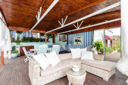 Un acogedor patio al aire libre, o como algunos dirían, un espacio acogedor, cuenta con un sofá de mimbre con cojines blancos, una mesa redonda, cojines a rayas en sillas de madera y plantas exuberantes. El espacio está cubierto con un techo de madera y tiene paneles de madera y azul claro para una relajación máxima.