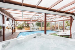 Una amplia zona de piscina cubierta, Espacios, cuenta con un techo de cristal y vigas de madera, con un jacuzzi en primer plano. Camastros, plantas exuberantes y sombrillas rodean la piscina, ofreciendo un ambiente relajante con una vista atractiva del exterior.