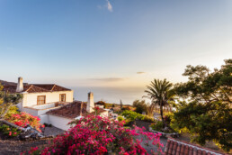 Un sereno paisaje costero al atardecer, con una casa de estilo español con techos de tejas rojas y buganvillas de un vibrante color rosa que envuelven los espacios. Árboles verdes y frondosos enmarcan la vista, mientras el océano se extiende más allá bajo un cielo despejado.