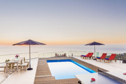 Una terraza en la azotea ofrece espacios con una pequeña piscina rectangular, rodeada de sillas rojas y sombrillas azules. A la izquierda hay una mesa de comedor de madera con sillas, perfecta para reuniones. La vista da al océano bajo un cielo de atardecer claro y colorido.