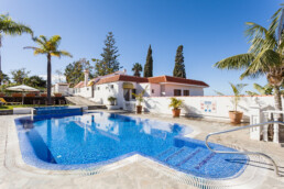 Un día soleado y luminoso en una villa con una gran y acogedora piscina rodeada de palmeras. Las paredes blancas y el techo de terracota de la villa se combinan a la perfección con la serenidad de los espacios. Las tumbonas con sombrillas salpican el fondo, realzando la atmósfera relajante.