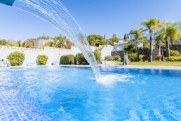 Una piscina de un azul resplandeciente con una cascada crea espacios acogedores en un entorno soleado al aire libre. Rodeada de sillas para descansar, palmeras y arbustos verdes, el cielo brillante y despejado realza la atmósfera veraniega.