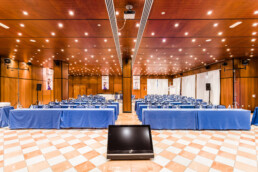 Espacios define la atmósfera de esta sala de conferencias, donde filas de mesas y sillas azules se enfrentan a un podio. El techo revestido de madera se ilumina con luces, mientras que las paredes están adornadas con pancartas. Una gran pantalla llama la atención en la parte delantera y el diseño de azulejos a cuadros añade un toque de elegancia a Espaicios.