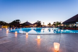 Una serena piscina al atardecer, rodeada de palmeras y suavemente iluminada por faroles brillantes, ofrece espacios tranquilos. El cielo pasa del azul al naranja pálido, con siluetas de estructuras con techo de paja al fondo.