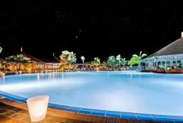 Una serena escena nocturna con una gran piscina iluminada rodeada de palmeras y acogedores edificios define estos encantadores espacios. El cielo está lleno de estrellas, una suave iluminación ambiental realza la atmósfera tranquila y una vela encendida se encuentra elegantemente en primer plano.