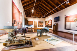 En una mesa de madera de Espacios se ha dispuesto con gran habilidad una exposición de museo que muestra equipos de impresión antiguos. La sala cuenta con paneles informativos que recubren las paredes, un techo de madera y una elegante y moderna lámpara. Una pantalla de televisión está montada estratégicamente en la pared derecha para ofrecer más información.