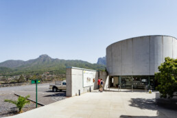 Un edificio moderno, cilíndrico y con grandes ventanales se alza sobre un paisaje montañoso y ofrece unas vistas impresionantes de los Espacios. Tres personas leen un cartel informativo en el exterior. Cerca, bajo el cielo azul claro, se ven una camioneta estacionada y un pequeño árbol.
