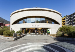 Un edificio moderno y circular con la etiqueta MVSEO se yergue orgulloso entre una exuberante vegetación. Grandes ventanales dejan entrar la luz a sus vibrantes espacios, mientras que un camino pavimentado invita a los visitantes a explorar sus maravillas. Todo esto tiene como fondo un cielo azul claro.