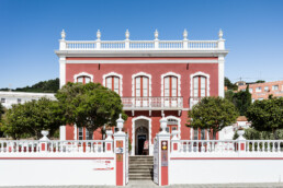 Un edificio rojo de dos pisos con molduras blancas y ventanas arqueadas, rodeado de frondosos árboles, ofrece espacios acogedores. Cuenta con una entrada cerrada con barandillas decorativas, todo bajo el abrazo de un cielo azul claro.