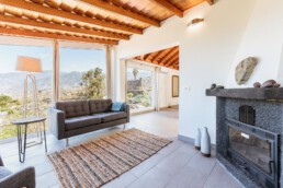 Una acogedora sala de estar con un sofá gris, una lámpara de pie y una alfombra tejida. Grandes ventanales ofrecen vistas panorámicas a la montaña. Una chimenea de piedra con decoración se alza en la esquina. La habitación cuenta con un techo de madera y azulejos de colores claros que crean un ambiente cálido.