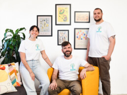 Tres personas con camisetas blancas a juego posan en un luminoso salón; dos de pie y una sentada en un sillón amarillo. La pared blanca que tienen detrás está decorada con láminas y pósters enmarcados.