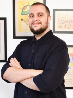 Un hombre con barba y camisa oscura abotonada está de pie con los brazos cruzados, sonriendo, delante de una pared con coloridas láminas de arte abstracto enmarcadas.