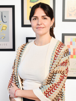 Una mujer con el pelo oscuro recogido en una coleta lleva un chal estampado sobre un top blanco, de pie con los brazos cruzados y sonriendo, delante de una pared con arte abstracto enmarcado de colores.