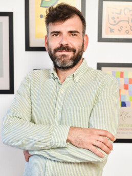 Un hombre con barba y pelo corto, vestido con una camisa de rayas verdes claras y blancas, está de pie con los brazos cruzados y sonríe a la cámara. En la pared detrás de él se ven coloridas obras de arte enmarcadas.