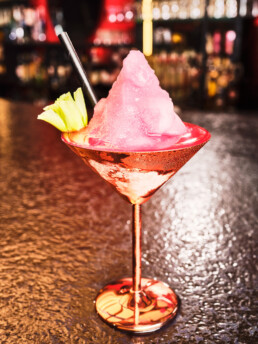Un cóctel helado de color rosa con un montón de hielo picado, adornado con una hoja de apio y servido en una copa de martini rosa metálico con pajita, reposa sobre una superficie texturizada en un bar poco iluminado.