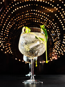Un gin-tonic con hielo, rodajas de lima y una varilla verde decorativa en un vaso globo, sobre un fondo oscuro con un patrón circular de luces brillantes.