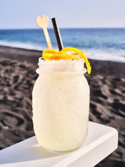 Un tarro de cristal lleno de una bebida helada de color amarillo claro, adornada con piel de limón, una pajita negra y un agitador de madera con forma de corazón, se encuentra en una repisa con una playa y el océano de fondo.