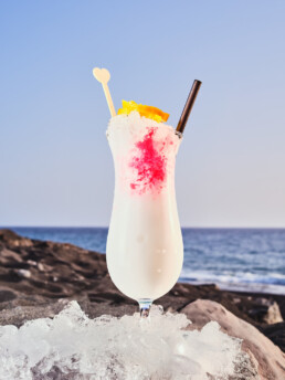 Una bebida tropical con líquido blanco, sirope rosa, rodaja de naranja, pajita y agitador de corazón se asienta sobre hielo frente a una playa rocosa y un océano azul bajo un cielo despejado.