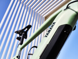 Primer plano de un cuadro de bicicleta Yamaha de color verde claro con una batería visible, sobre una moderna estructura arquitectónica con vigas verticales blancas y un cielo azul despejado.
