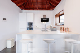 Cocina moderna con armarios blancos, isla y tres taburetes de bar blancos. Electrodomésticos de acero inoxidable, botes de cobre y una gran ventana. La sala cuenta con un alto techo abovedado de madera.